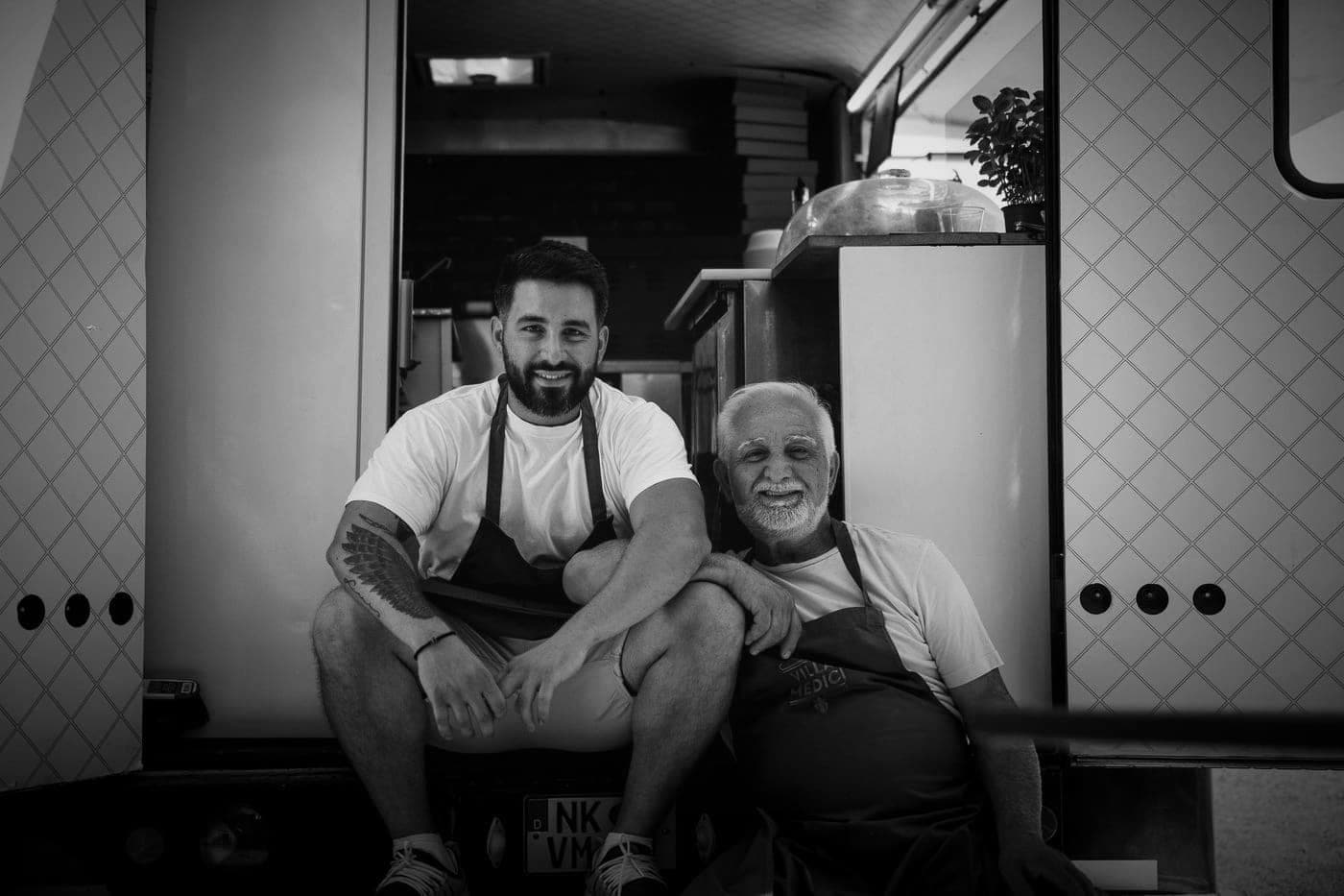 Zwei Männer in Schürzen sitzen nebeneinander in der Durchreiche des Foodtrucks