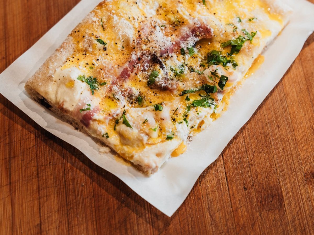 Ein Stück Pinsa al taglio mit Ei, Speck, Kräutern und geriebenem Käse auf Backpapier