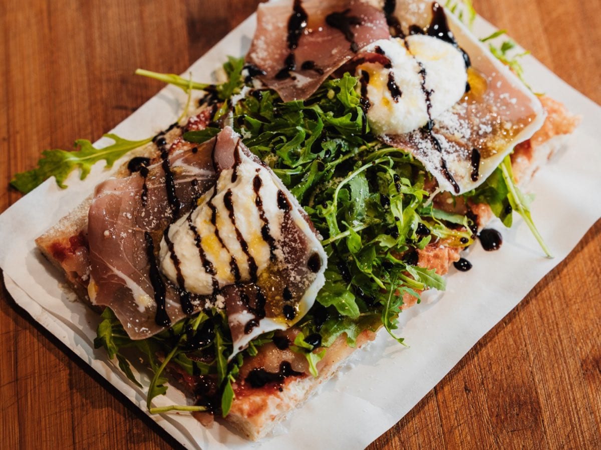 Ein Stück Pinsa al taglio mit Rohschinken, Burrata, Rucola und Balsamico auf Backpapier